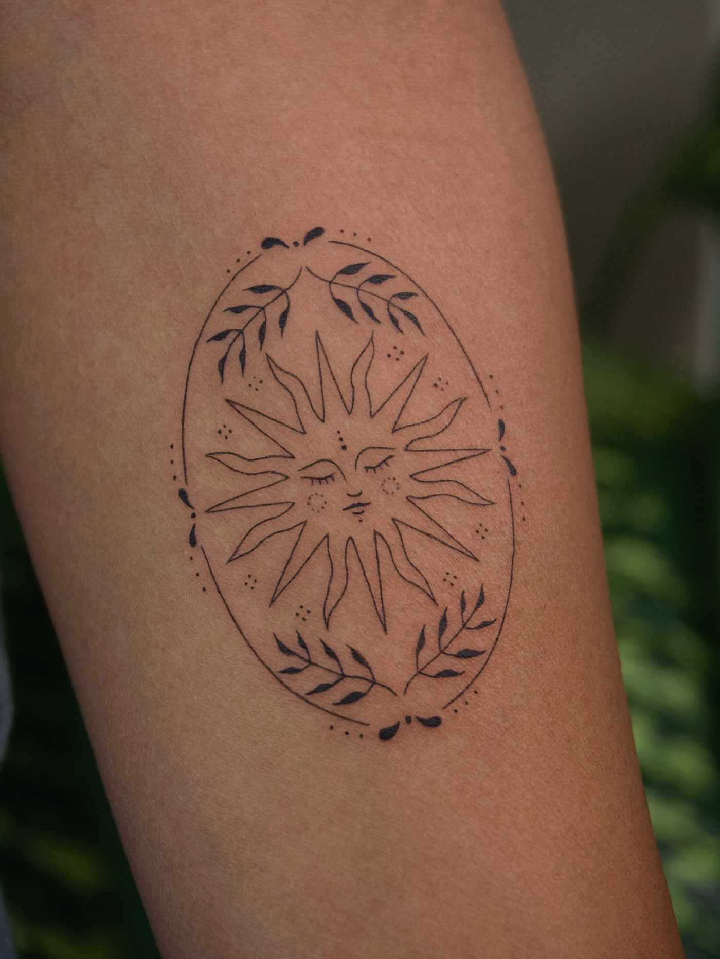 Minimalist tattoo of a sun with a face inside a leaf-framed oval, arm