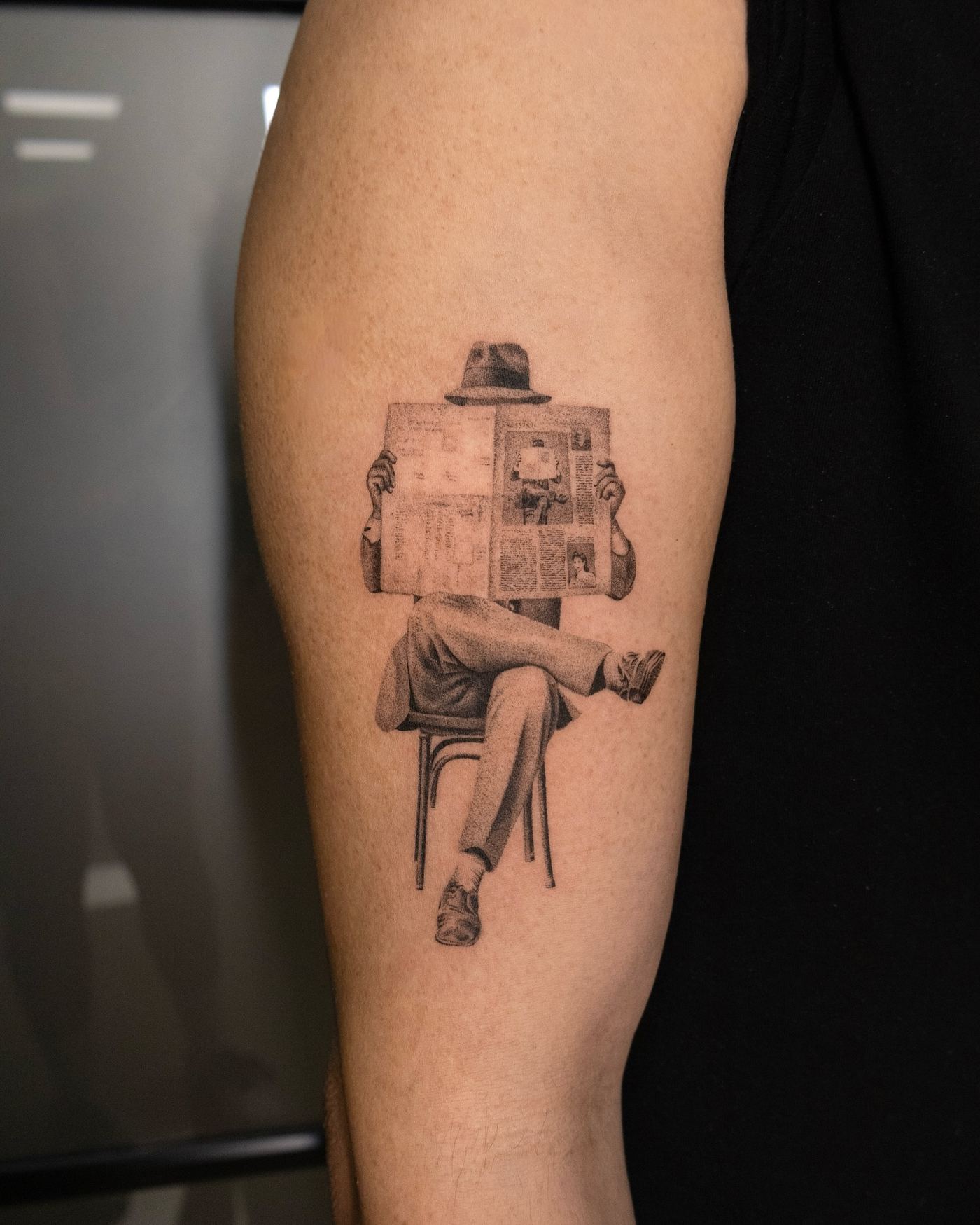 Micro-realistic tattoo of a seated figure reading the newspaper, forearm